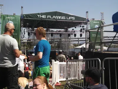 Sunny outdoor concert stage for The Flaming Lips with crowd relaxing on grass nearby.